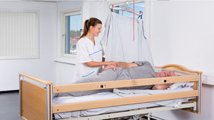 A care taker turning an old man with WendyLett4Way Sliding DrawSheet  attached to a hoist on the ceiling