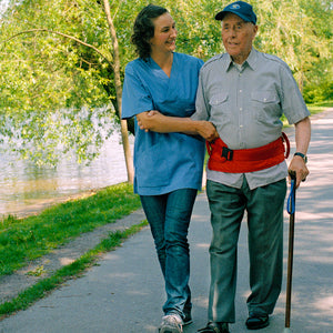 Direct Healthcare Group FlexiBelt Sit-to-Stand and Walking Support Belt worn by senior while being assisted by a caregiver during a morning walk beside a river