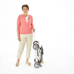 Motion Healthcare mLite, 4 Wheel, Lightweight, Folding Electric Mobility Scooter, grey Lithium Battery woman standing next to scooter holding the extending handle